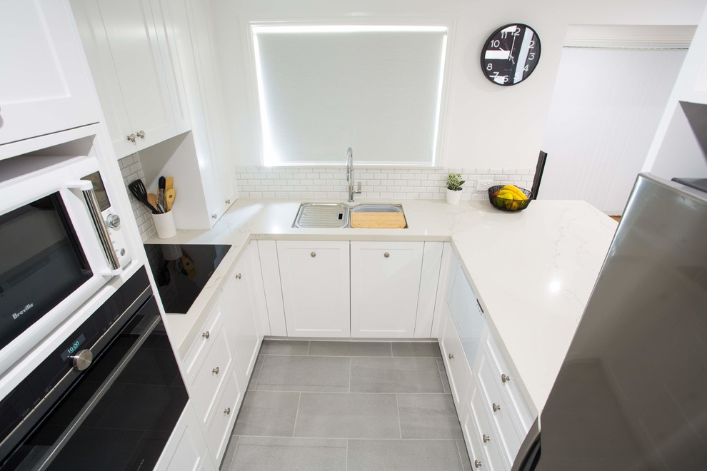 Photo of a small traditional u-shaped kitchen pantry in Brisbane with a double-bowl sink, shaker cabinets, white cabinets, quartz benchtops, grey splashback, ceramic splashback, black appliances, porcelain floors, with island, grey floor and white benchtop.
