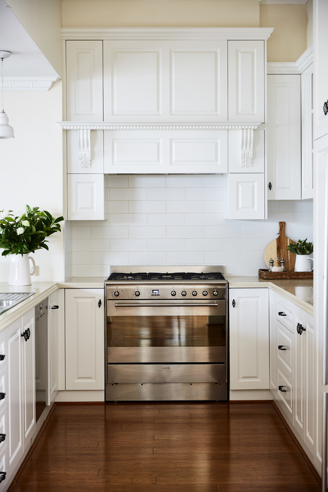 This is an example of a country u-shaped eat-in kitchen in Melbourne with a double-bowl sink, recessed-panel cabinets, white cabinets, quartz benchtops, white splashback, subway tile splashback, stainless steel appliances, bamboo floors, brown floor and beige benchtop.