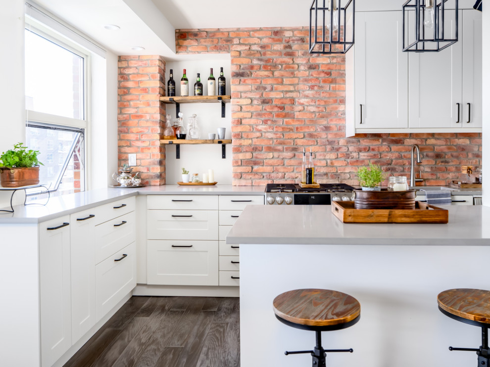 This is an example of a small industrial l-shaped kitchen/diner in New York with a submerged sink, shaker cabinets, white cabinets, red splashback, brick splashback, stainless steel appliances, grey worktops, laminate floors, a breakfast bar and grey floors.