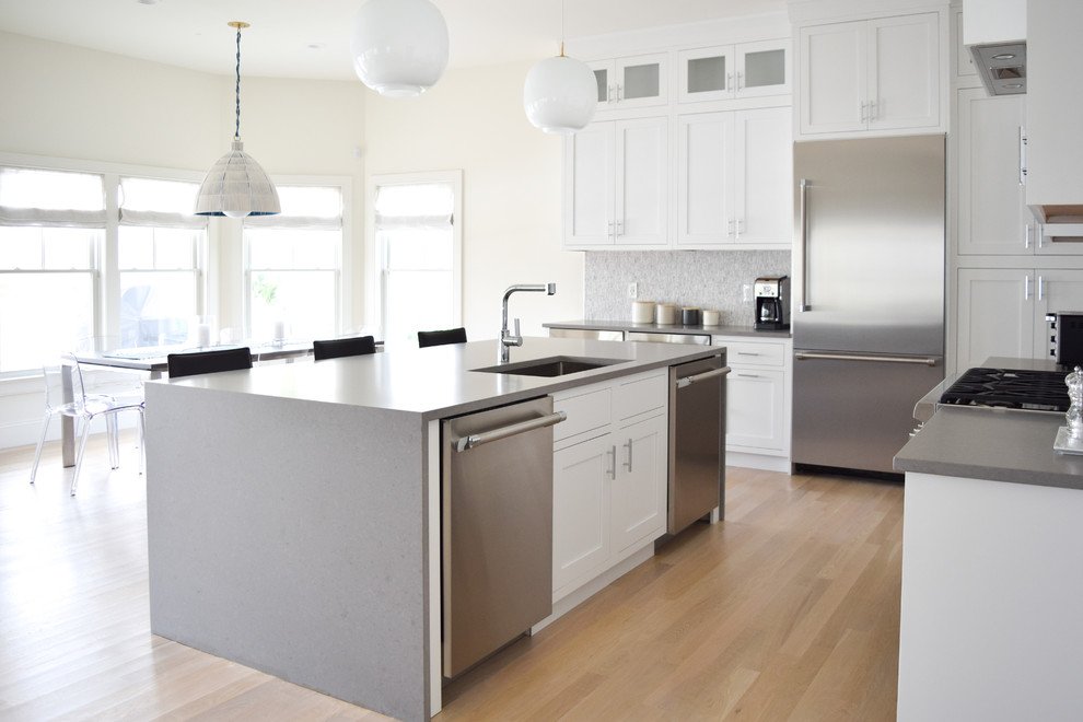 Inspiration for a mid-sized beach style l-shaped eat-in kitchen in New York with an undermount sink, recessed-panel cabinets, white cabinets, quartz benchtops, grey splashback, marble splashback, stainless steel appliances, light hardwood floors and with island.