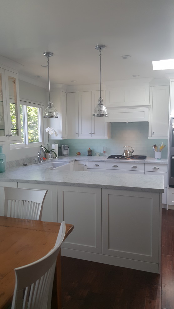 Mid-sized beach style u-shaped eat-in kitchen in Los Angeles with a farmhouse sink, shaker cabinets, white cabinets, marble benchtops, blue splashback, glass tile splashback, stainless steel appliances, dark hardwood floors, a peninsula and brown floor.