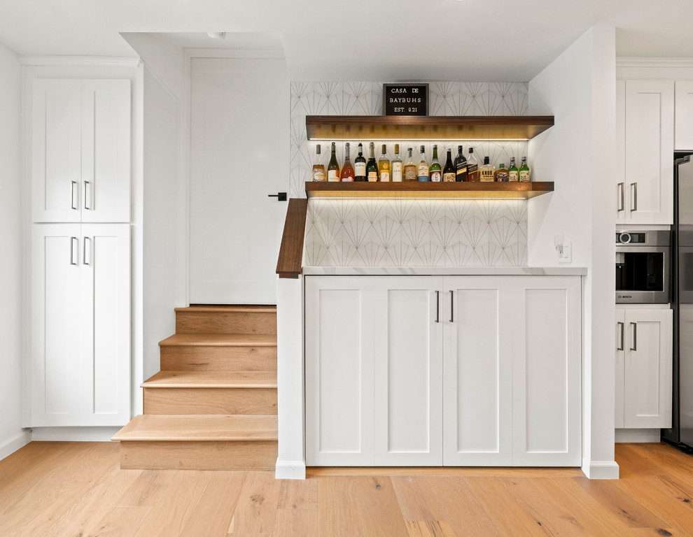 Inspiration for a mid-sized transitional home bar in Los Angeles with shaker cabinets, white cabinets, quartz benchtops, white splashback, cement tile splashback, light hardwood floors, brown floor and white benchtop.