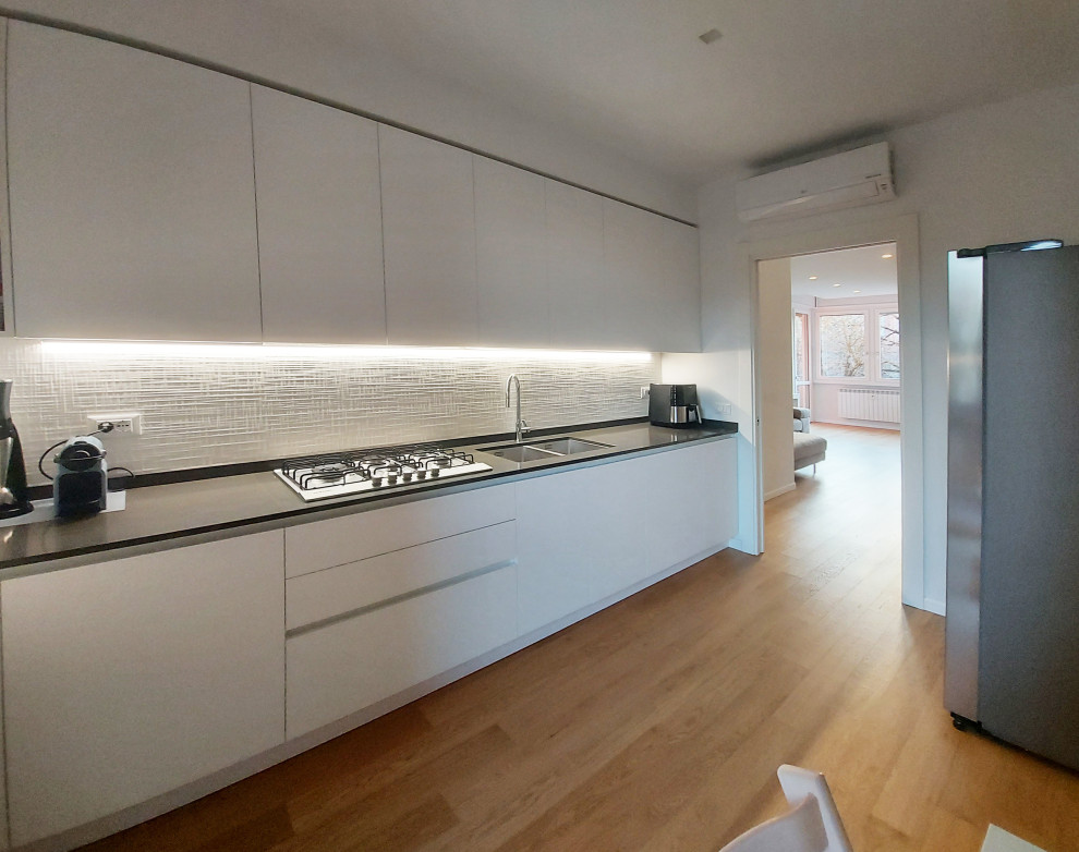 Design ideas for a mid-sized contemporary galley separate kitchen in Milan with a drop-in sink, flat-panel cabinets, white cabinets, quartzite benchtops, white splashback, porcelain splashback, light hardwood floors, black benchtop and recessed.