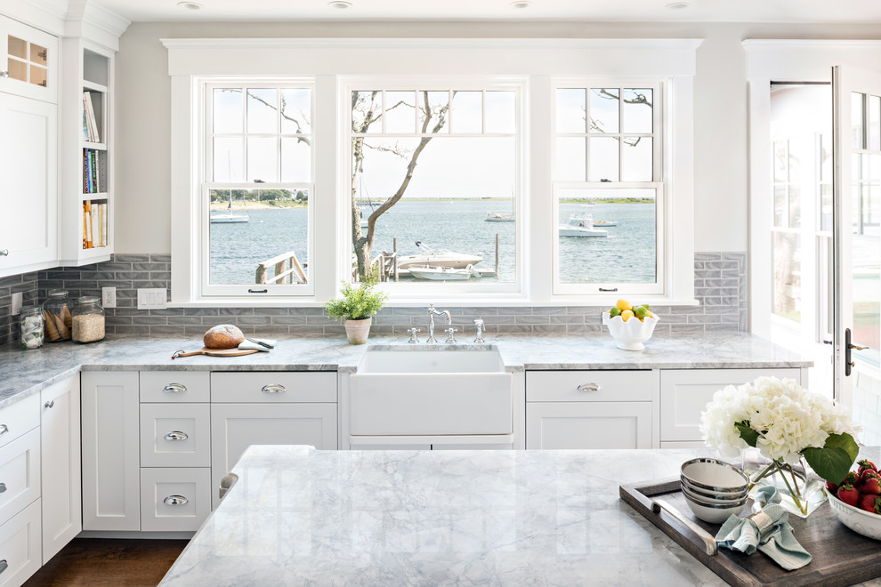 Design ideas for a beach style kitchen in Boston with a farmhouse sink, shaker cabinets, white cabinets, quartzite benchtops, grey splashback, porcelain splashback, stainless steel appliances and medium hardwood floors.