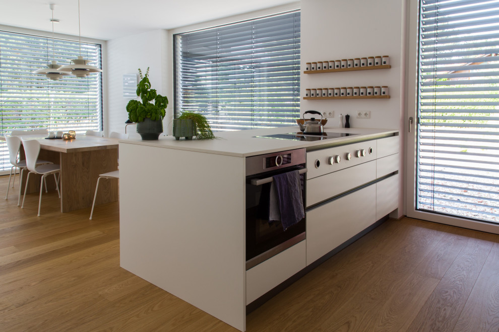 На фото: серо-белая кухня-гостиная в современном стиле с белыми фасадами, белой столешницей, плоскими фасадами, паркетным полом среднего тона и полуостровом с