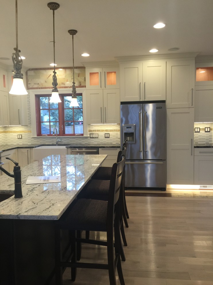 Eat-in kitchen - mid-sized transitional u-shaped light wood floor and beige floor eat-in kitchen idea in Boston with recessed-panel cabinets, white cabinets, stainless steel appliances, an island, a farmhouse sink, granite countertops, beige backsplash, stone tile backsplash and multicolored countertops