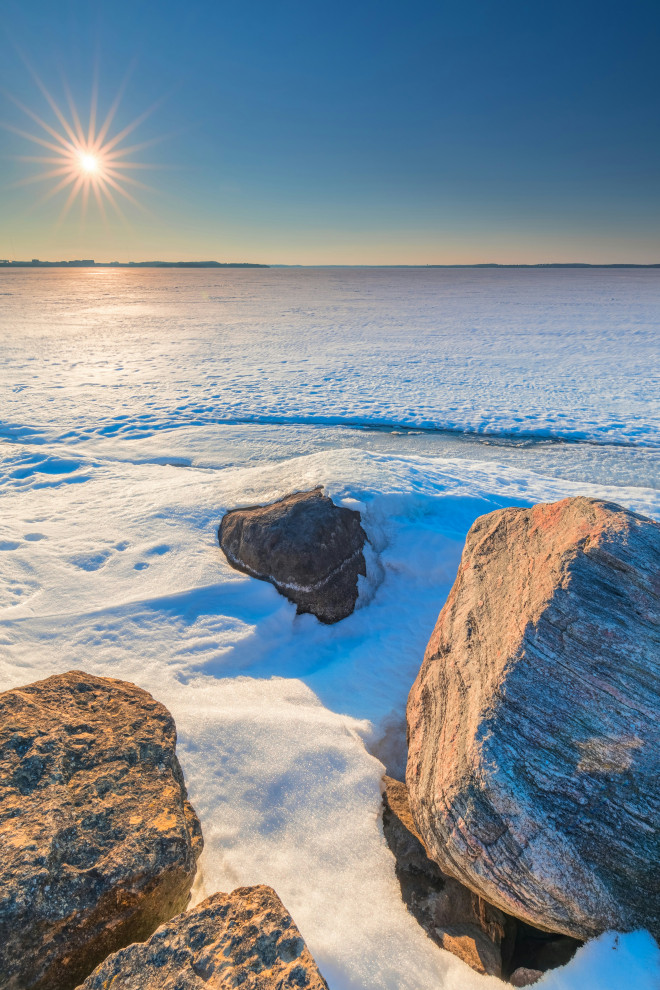 rocks at shore of frozen snowy wisconsin lake with sunburst in blue sky