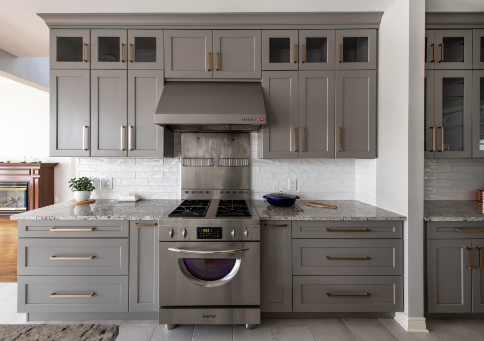Example of a mid-sized transitional l-shaped porcelain tile and gray floor open concept kitchen design in Ottawa with a farmhouse sink, shaker cabinets, gray cabinets, granite countertops, white backsplash, ceramic backsplash, stainless steel appliances, an island and multicolored countertops