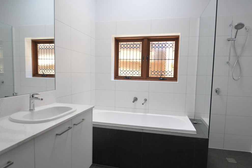 This is an example of a mid-sized contemporary kids bathroom in Melbourne with flat-panel cabinets, white cabinets, a drop-in tub, an open shower, a one-piece toilet, white tile, porcelain tile, white walls, porcelain floors, a drop-in sink, solid surface benchtops, grey floor and an open shower.