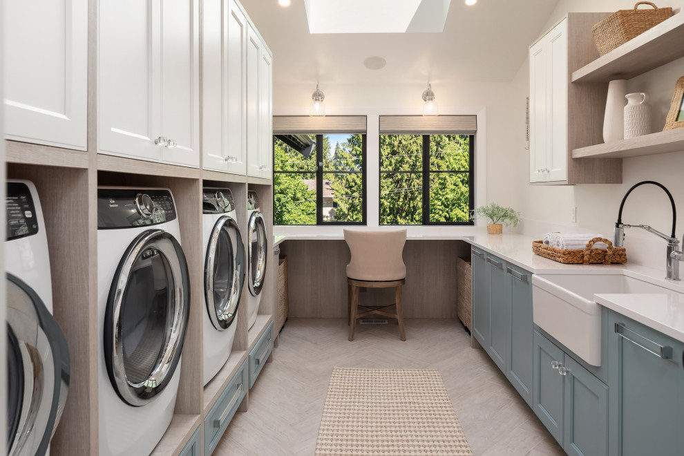 Large transitional laundry room in Seattle with a farmhouse sink, shaker cabinets, blue cabinets, quartz benchtops, stone slab splashback, white walls, porcelain floors, a side-by-side washer and dryer, grey floor and white benchtop.