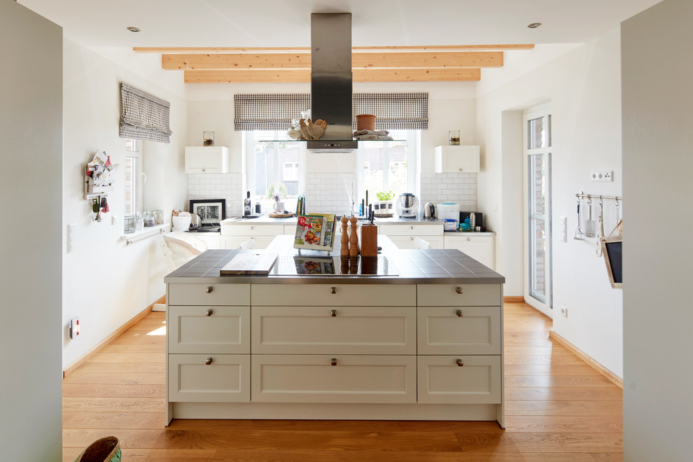This is an example of a mid-sized country galley open plan kitchen in Bremen with shaker cabinets, white cabinets, tile benchtops, white splashback, subway tile splashback, medium hardwood floors, with island, brown floor and grey benchtop.