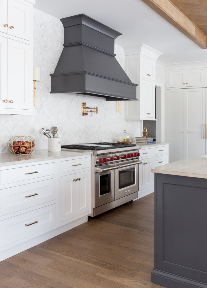 Transitional l-shaped kitchen in New York with a farmhouse sink, shaker cabinets, white cabinets, white splashback, stainless steel appliances, medium hardwood floors, with island, brown floor and beige benchtop.