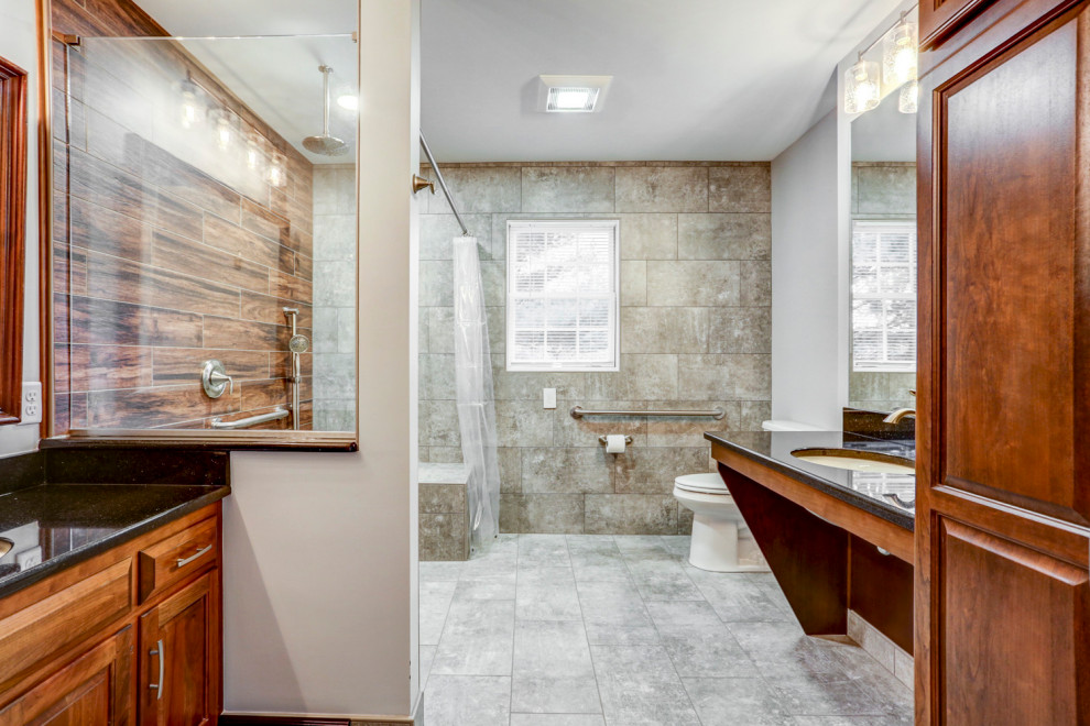 Photo of a large transitional master bathroom in Other with raised-panel cabinets, brown cabinets, an open shower, a two-piece toilet, gray tile, ceramic tile, grey walls, ceramic floors, an undermount sink, granite benchtops, grey floor, an open shower, black benchtops, a shower seat, a double vanity and a built-in vanity.