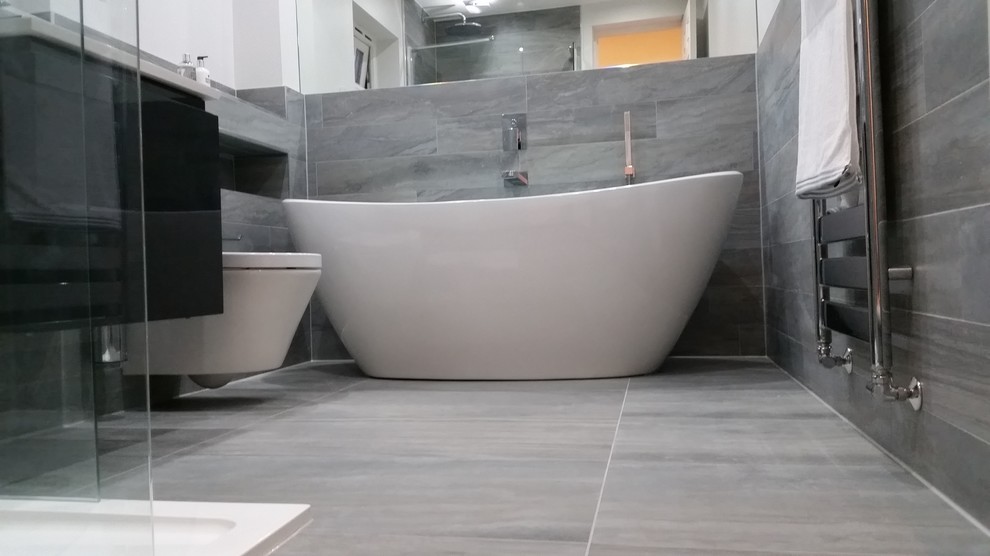 This is an example of a mid-sized modern bathroom in Hertfordshire with a wall-mount sink, a freestanding tub, an open shower, a wall-mount toilet, gray tile, porcelain tile, white walls and porcelain floors.