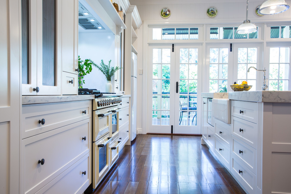 This is an example of a large traditional galley eat-in kitchen in Brisbane with shaker cabinets, white cabinets, white splashback, with island, a farmhouse sink, subway tile splashback, dark hardwood floors, white appliances and marble benchtops.