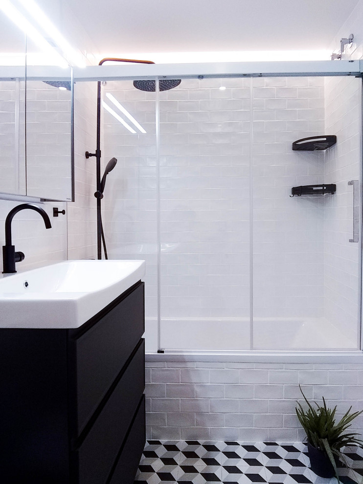 This is an example of a medium sized modern grey and white ensuite bathroom in Barcelona with raised-panel cabinets, black cabinets, a built-in bath, a shower/bath combination, a wall mounted toilet, white tiles, ceramic tiles, white walls, cement flooring, an integrated sink, solid surface worktops, black floors, a sliding door, white worktops, an enclosed toilet, a single sink and a built in vanity unit.