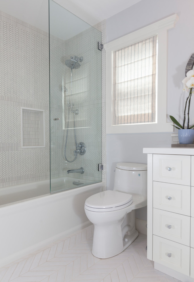 Photo of a mid-sized beach style kids bathroom in Huntington with shaker cabinets, white cabinets, a drop-in tub, a shower/bathtub combo, a one-piece toilet, gray tile, ceramic tile, grey walls, ceramic floors, a drop-in sink, quartzite benchtops, white floor, a hinged shower door and white benchtops.