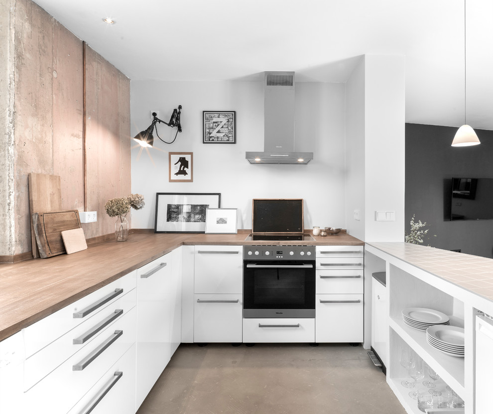 Photo of a small industrial u-shaped open plan kitchen in Hamburg with white cabinets, wood benchtops, a peninsula, grey floor, brown benchtop, flat-panel cabinets, black appliances and concrete floors.
