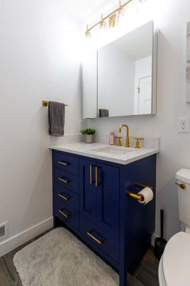 Пример оригинального дизайна: ванная комната среднего размера в стиле неоклассика (современная классика) с фасадами с утопленной филенкой, синими фасадами, ванной в нише, душем над ванной, раздельным унитазом, серой плиткой, керамической плиткой, белыми стенами, полом из винила, душевой кабиной, врезной раковиной, столешницей из искусственного кварца, коричневым полом, душем с раздвижными дверями, белой столешницей, тумбой под одну раковину и встроенной тумбой