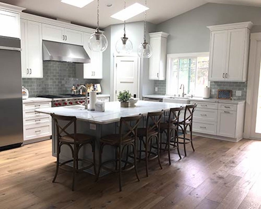 Inspiration for a mid-sized transitional l-shaped open plan kitchen in New York with a farmhouse sink, shaker cabinets, white cabinets, quartzite benchtops, grey splashback, subway tile splashback, stainless steel appliances, medium hardwood floors, with island and brown floor.