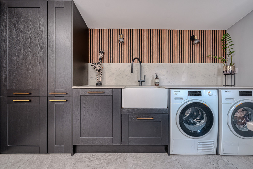 ロンドンにあるお手頃価格の広いコンテンポラリースタイルのおしゃれなランドリールーム (L型、エプロンフロントシンク、シェーカースタイル扉のキャビネット、黒いキャビネット、珪岩カウンター、白いキッチンパネル、クオーツストーンのキッチンパネル、グレーの壁、磁器タイルの床、左右配置の洗濯機・乾燥機、グレーの床、白いキッチンカウンター、折り上げ天井、パネル壁) の写真