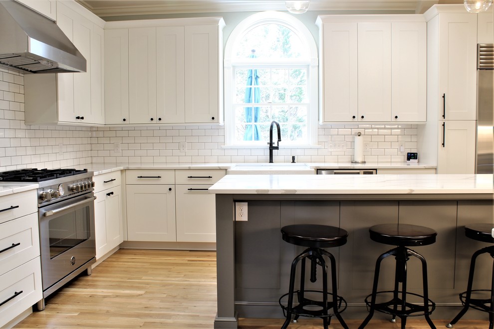 Large transitional galley light wood floor eat-in kitchen photo with a farmhouse sink, shaker cabinets, white cabinets, quartz countertops, white backsplash, ceramic backsplash, stainless steel appliances, an island and white countertops