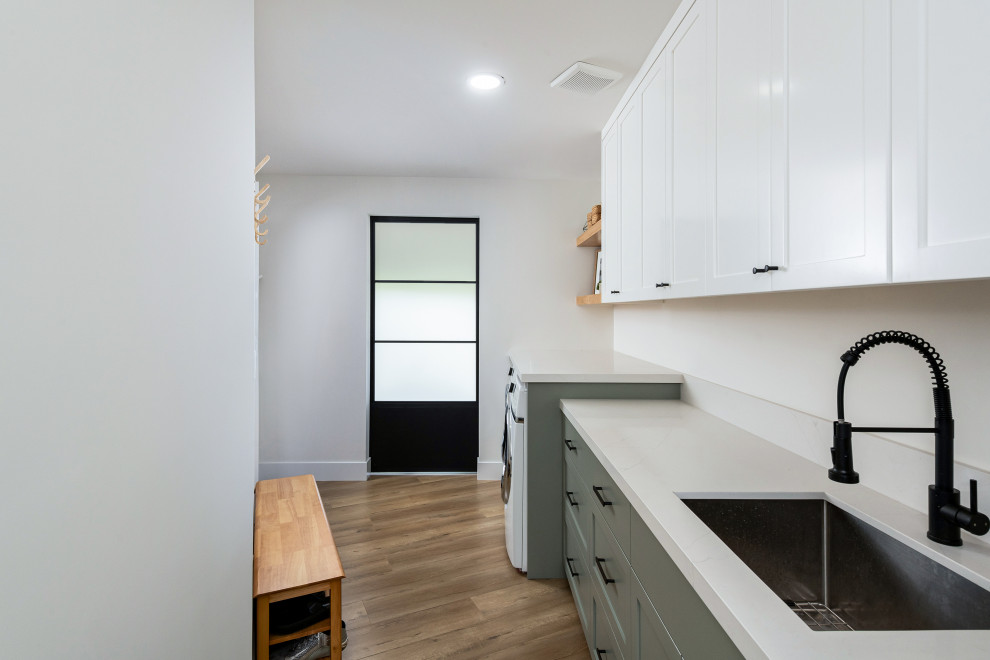 Design ideas for a mid-sized beach style dedicated laundry room in Orange County with a drop-in sink, green cabinets, quartz benchtops, white splashback, engineered quartz splashback, white walls, medium hardwood floors, a side-by-side washer and dryer and white benchtop.