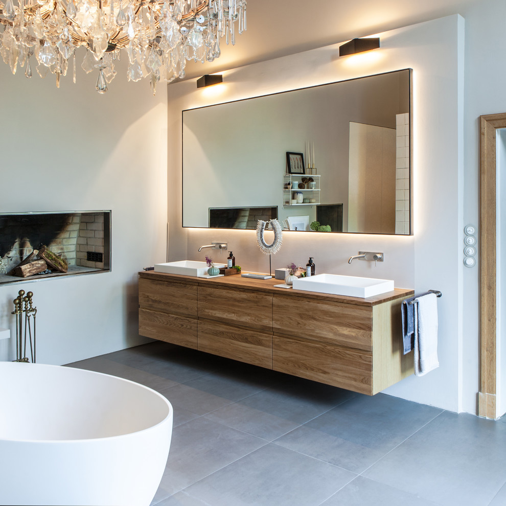 Photo of a large contemporary master bathroom in Hamburg with flat-panel cabinets, light wood cabinets, a freestanding tub, a curbless shower, a wall-mount toilet, white tile, limestone, beige walls, ceramic floors, a drop-in sink, solid surface benchtops, grey floor, an open shower and brown benchtops.
