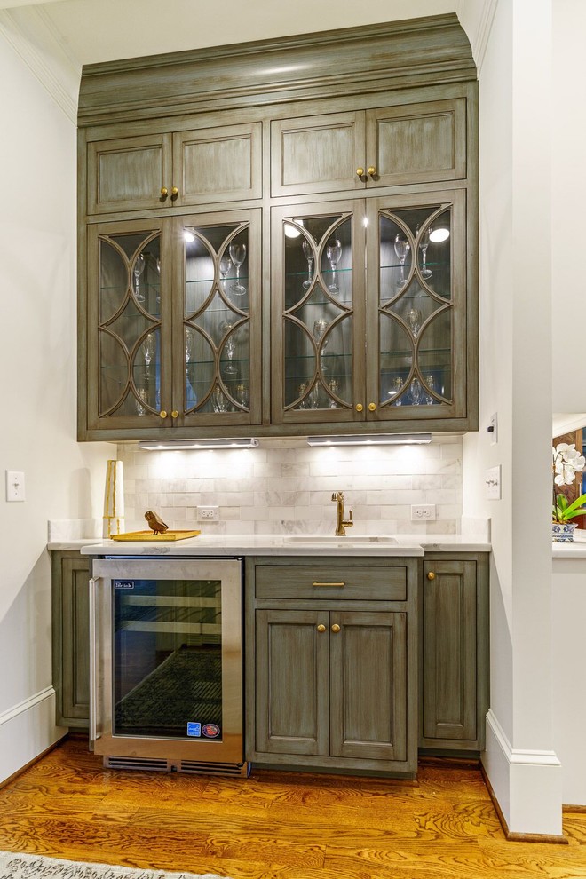 Photo of a mid-sized traditional single-wall wet bar in Other with an undermount sink, recessed-panel cabinets, distressed cabinets, marble benchtops, grey splashback, marble splashback, light hardwood floors, brown floor and grey benchtop.