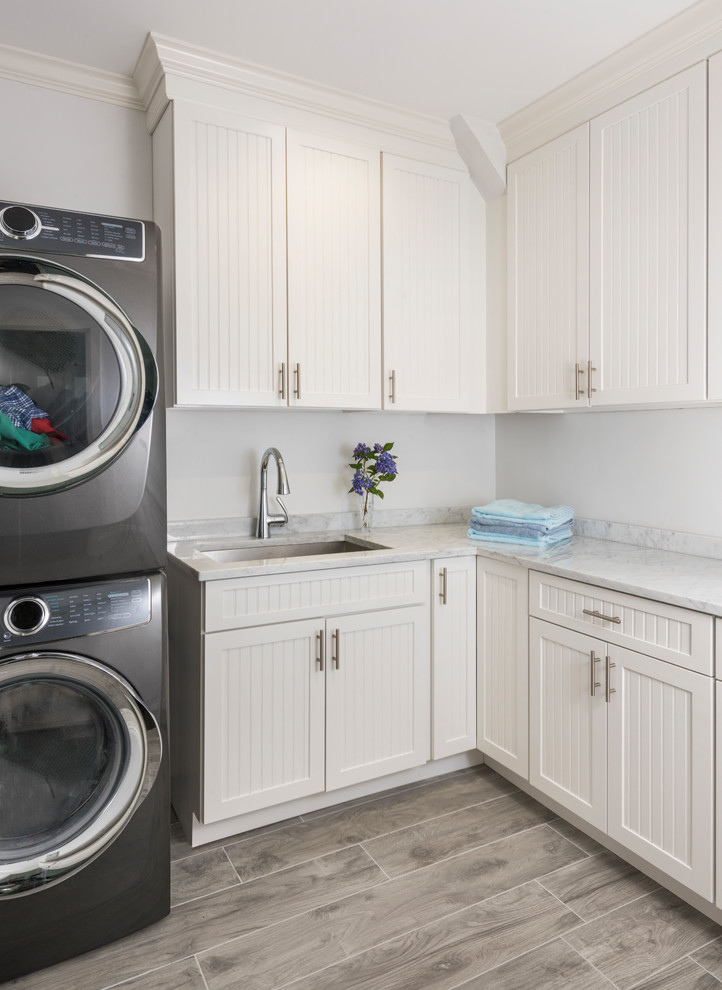 This is an example of a mid-sized transitional l-shaped dedicated laundry room in Boston with an undermount sink, beaded inset cabinets, white cabinets, marble benchtops, grey walls, ceramic floors, a stacked washer and dryer and grey floor.