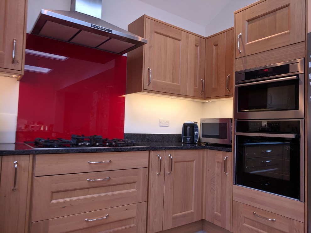 Inspiration for a large traditional u-shaped open plan kitchen in London with a double-bowl sink, shaker cabinets, brown cabinets, granite benchtops, red splashback, glass sheet splashback, black appliances, a peninsula and grey floor.