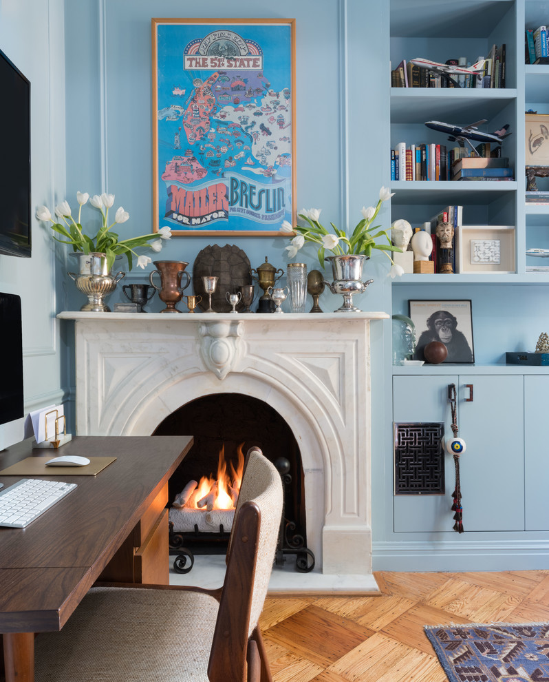 This is an example of a small eclectic home office in New York with a library, blue walls, medium hardwood floors, a standard fireplace, a stone fireplace surround, a freestanding desk and brown floor.