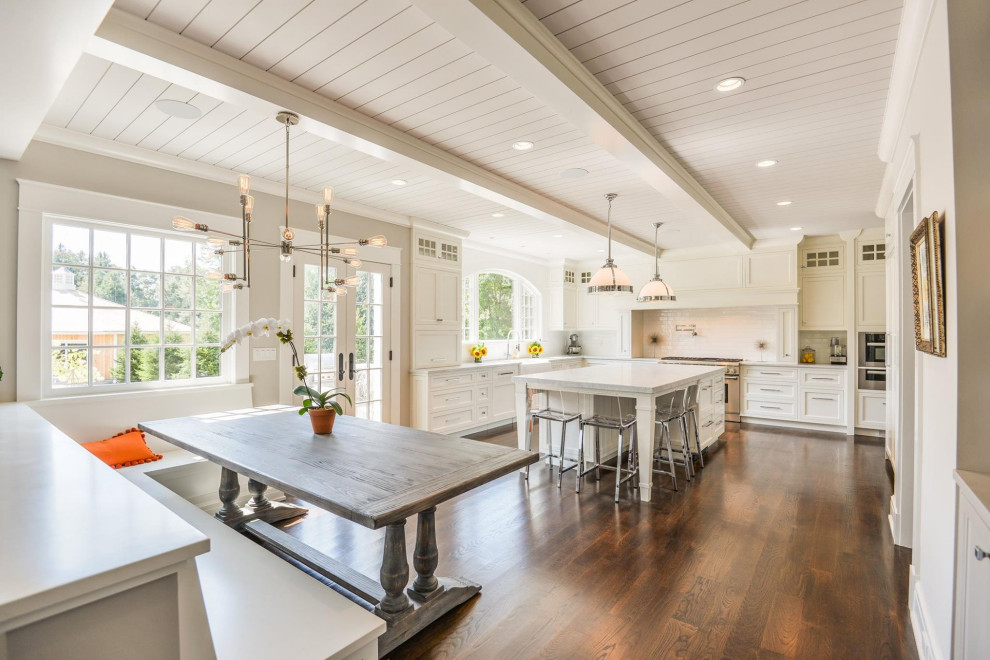 Photo of a traditional l-shaped eat-in kitchen in New York with a farmhouse sink, recessed-panel cabinets, white cabinets, white splashback, stainless steel appliances, dark hardwood floors, with island, brown floor, white benchtop, exposed beam and timber.