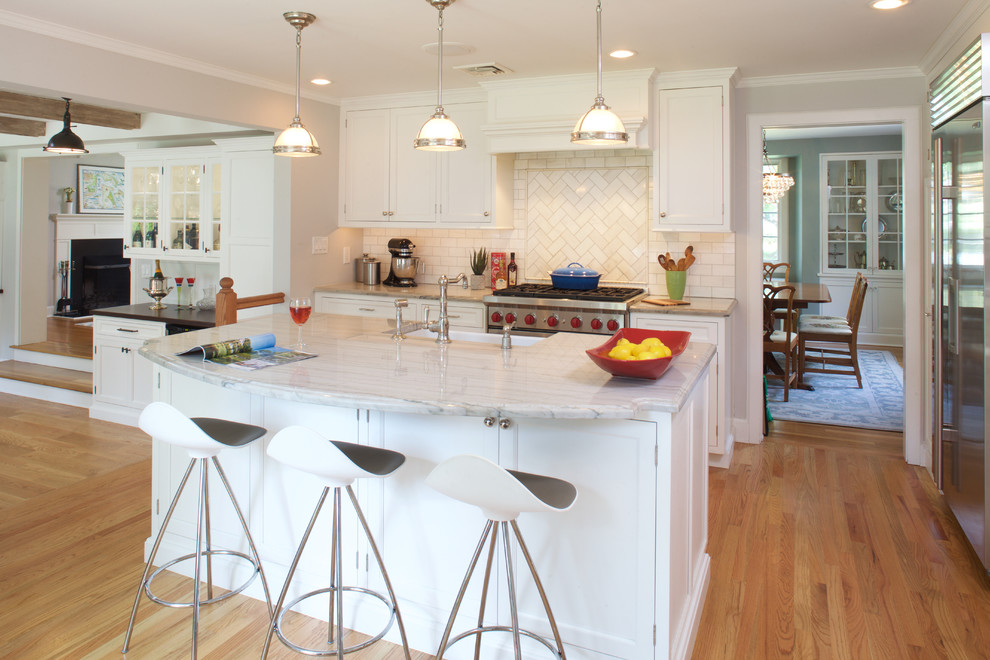 Photo of a mid-sized transitional l-shaped kitchen in New York with a farmhouse sink, recessed-panel cabinets, white cabinets, white splashback, subway tile splashback, stainless steel appliances, light hardwood floors and with island.