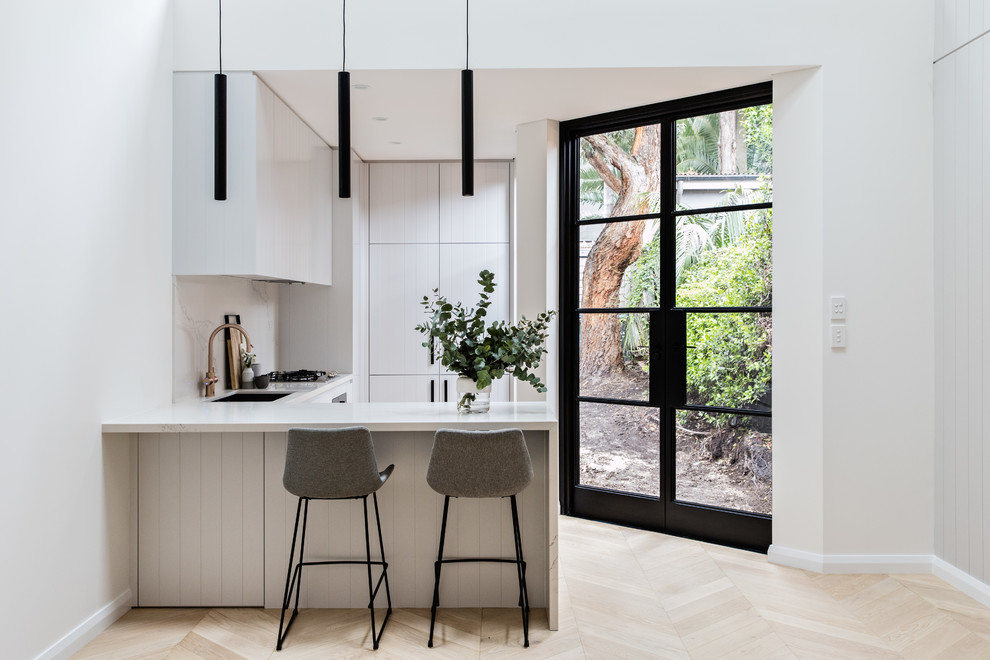 This is an example of a contemporary l-shaped kitchen in Sydney with an undermount sink, flat-panel cabinets, grey cabinets, white splashback, marble splashback, light hardwood floors, a peninsula, beige floor and white benchtop.