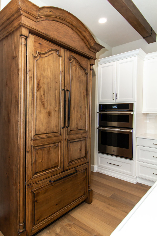 Inredning av ett stort vit vitt kök, med en undermonterad diskho, luckor med infälld panel, vita skåp, bänkskiva i kvarts, vitt stänkskydd, stänkskydd i keramik, rostfria vitvaror, mellanmörkt trägolv, en köksö och brunt golv