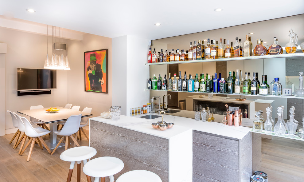 This is an example of a contemporary seated home bar in London with an undermount sink, flat-panel cabinets, light wood cabinets, light hardwood floors and white benchtop.