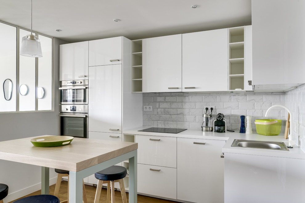 Mid-sized contemporary l-shaped open plan kitchen in Paris with white splashback, subway tile splashback, light hardwood floors, a single-bowl sink, flat-panel cabinets, white cabinets, stainless steel appliances and with island.