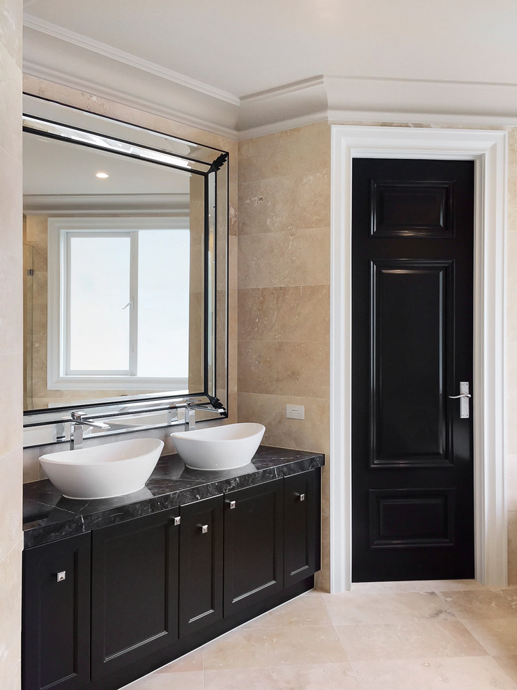Large transitional master bathroom in Melbourne with shaker cabinets, black cabinets, a freestanding tub, a double shower, a wall-mount toilet, beige tile, travertine, beige walls, travertine floors, a console sink, marble benchtops, beige floor, a hinged shower door and black benchtops.