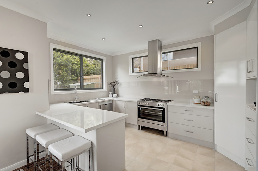 Small modern u-shaped open plan kitchen in Melbourne with a double-bowl sink, flat-panel cabinets, white cabinets, solid surface benchtops, stainless steel appliances, light hardwood floors, a peninsula and glass sheet splashback.