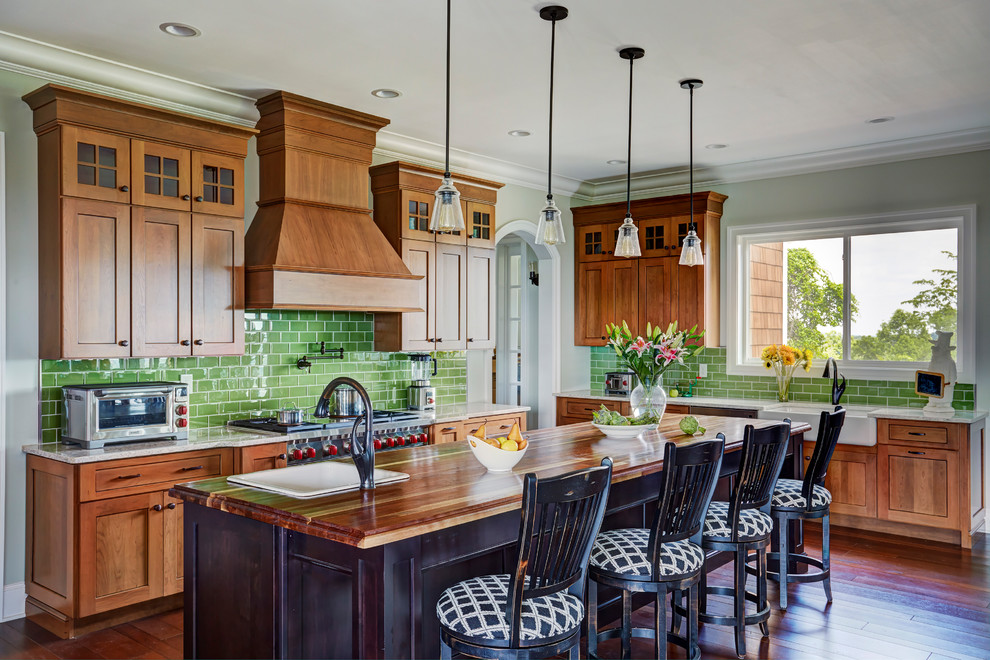 Photo of an arts and crafts l-shaped kitchen in Huntington with a farmhouse sink, shaker cabinets, medium wood cabinets, wood benchtops, green splashback, subway tile splashback, stainless steel appliances, dark hardwood floors, with island, brown floor and brown benchtop.