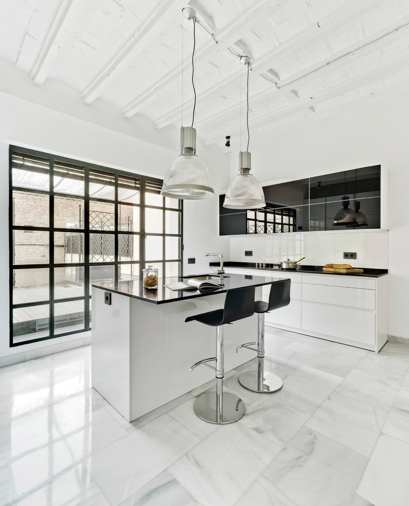 This is an example of a contemporary single-wall kitchen in Madrid with an undermount sink, flat-panel cabinets, granite benchtops, white splashback, black appliances, with island, white floor, marble floors, glass sheet splashback and black benchtop.