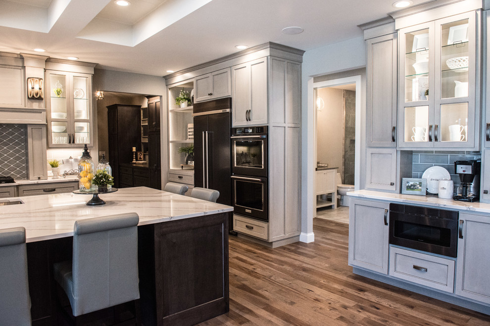Inspiration for a huge transitional l-shaped medium tone wood floor and brown floor open concept kitchen remodel with a single-bowl sink, glass-front cabinets, gray cabinets, quartz countertops, gray backsplash, ceramic backsplash, stainless steel appliances, an island and white countertops