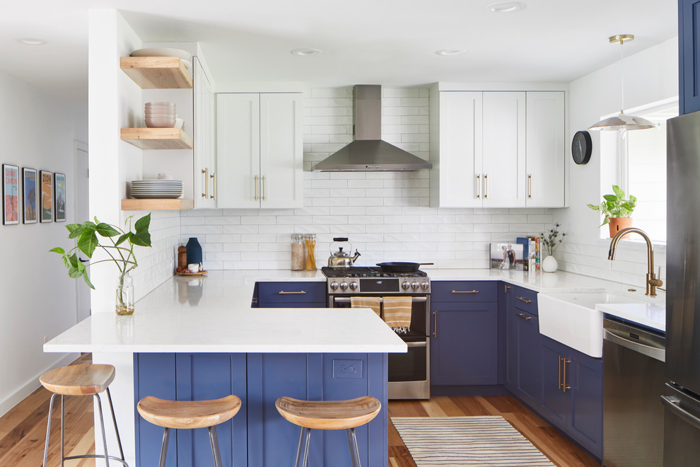 Design ideas for a mid-sized transitional u-shaped eat-in kitchen in Austin with a farmhouse sink, shaker cabinets, blue cabinets, quartz benchtops, white splashback, ceramic splashback, stainless steel appliances, a peninsula, brown floor, white benchtop and light hardwood floors.