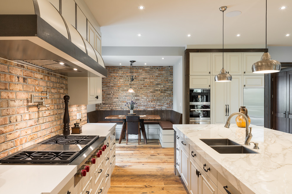 Design ideas for an industrial l-shaped open plan kitchen in Toronto with a double-bowl sink, beaded inset cabinets, white cabinets, brick splashback, stainless steel appliances, with island and beige floor.