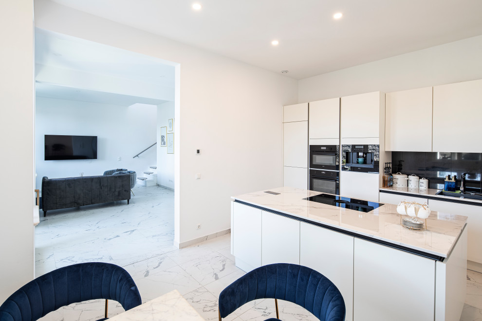 Mid-sized contemporary single-wall open plan kitchen in Berlin with a double-bowl sink, flat-panel cabinets, white cabinets, marble benchtops, black splashback, granite splashback, black appliances, marble floors, with island, white floor and white benchtop.