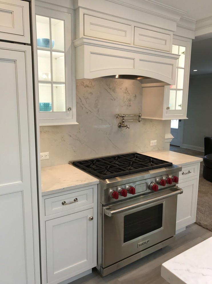Mid-sized transitional u-shaped light wood floor and brown floor open concept kitchen photo with a farmhouse sink, recessed-panel cabinets, white cabinets, marble countertops, white backsplash, marble backsplash, stainless steel appliances and an island