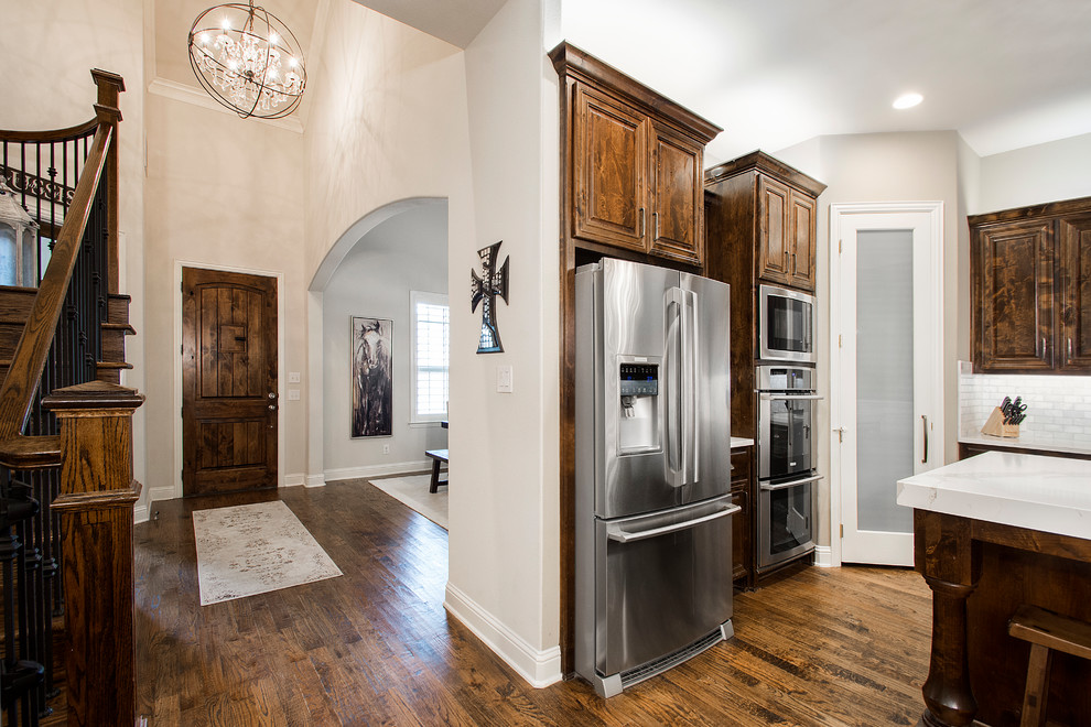 Photo of a large transitional u-shaped eat-in kitchen in Dallas with a farmhouse sink, raised-panel cabinets, dark wood cabinets, quartz benchtops, grey splashback, marble splashback, stainless steel appliances, medium hardwood floors, with island, brown floor and white benchtop.