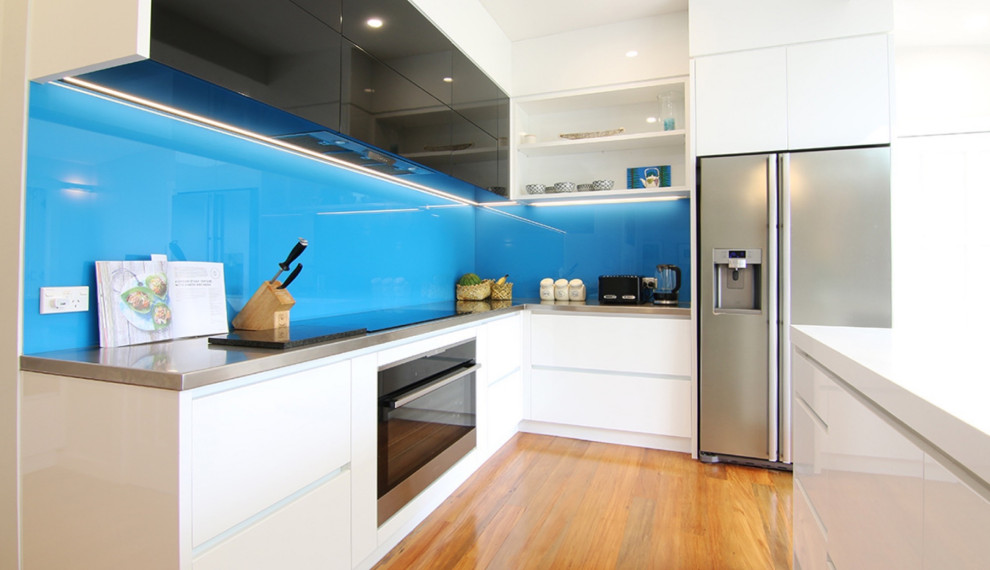 Photo of a mid-sized contemporary l-shaped eat-in kitchen in Sydney with raised-panel cabinets, white cabinets, solid surface benchtops, blue splashback, glass sheet splashback, stainless steel appliances, light hardwood floors, with island and white benchtop.