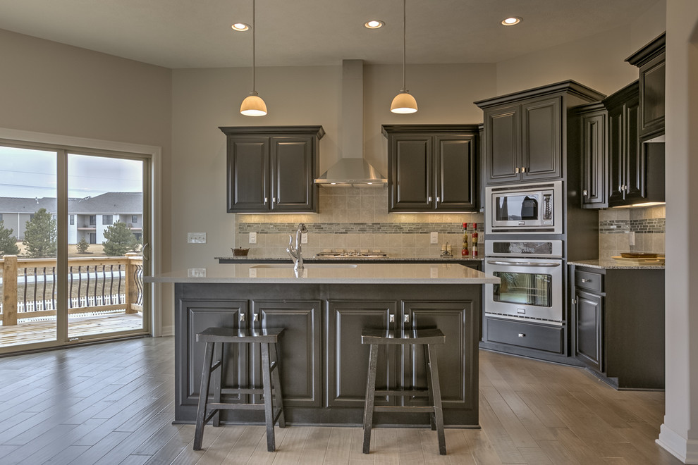 Photo of a mid-sized modern single-wall open plan kitchen in Omaha with a double-bowl sink, raised-panel cabinets, brown cabinets, granite benchtops, beige splashback, ceramic splashback, stainless steel appliances, light hardwood floors and with island.